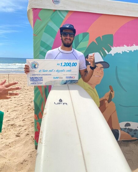 A person holding a check for R$ 1,200.00, standing next to a surfboard on a beach with a colorful mural in the background. 2