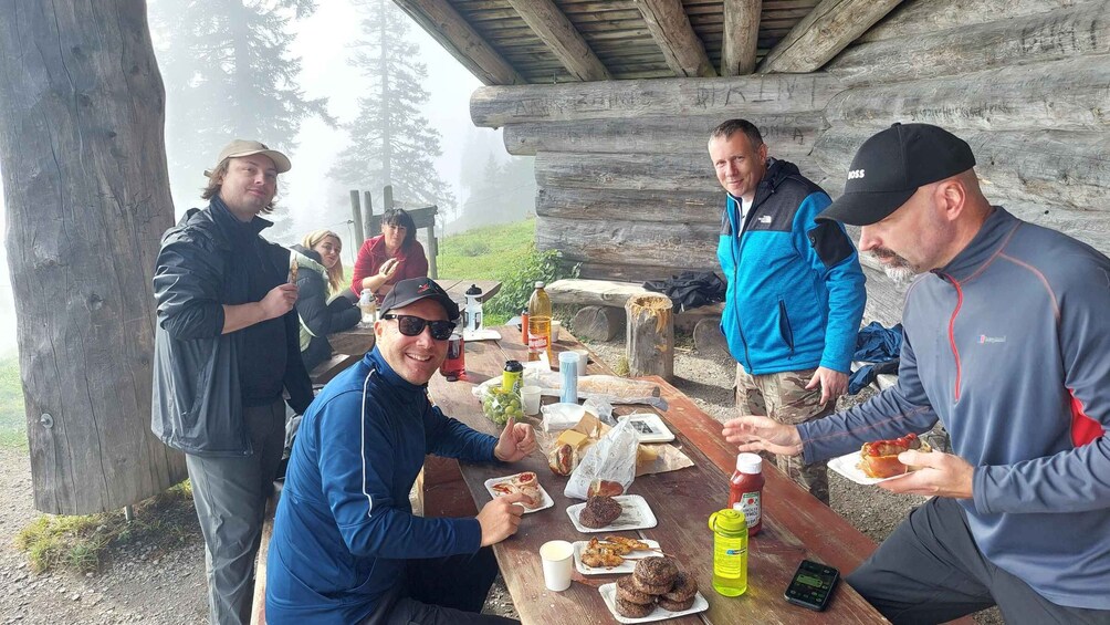 A group of people gathered around a wooden picnic table outdoors, with food and drinks on the table. 2