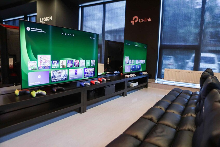 A gaming lounge with two large screens displaying a video game interface, a black leather couch, and gaming controllers. 3