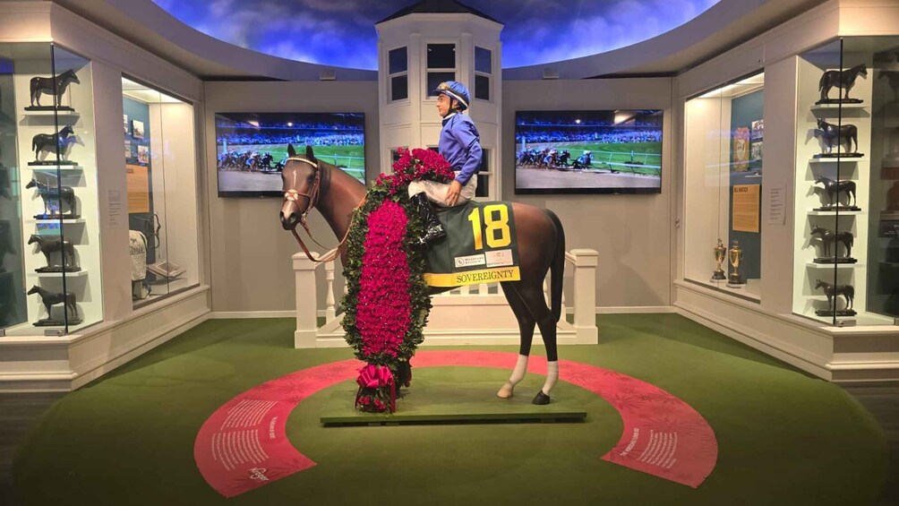 A jockey on a horse with a number 18 blanket, surrounded by horse statues and trophies. 2