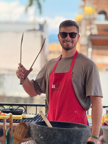 A person wearing a red apron holding cooking utensils, standing in front of a grill with food items. 5