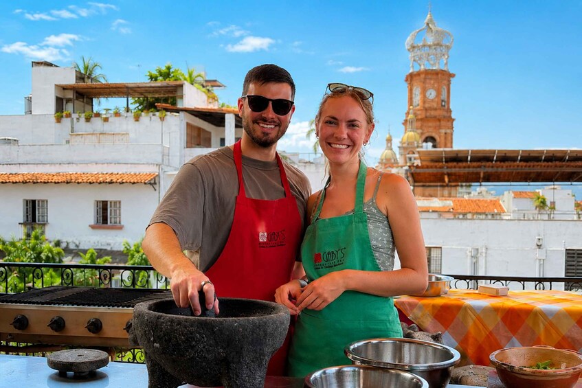 Two people standing by a grill with a stone mortar and pestle, a metal bowl, and a table with a checkered tablecloth. In the background, there is a building with a clock tower and a clear blue sky. 3