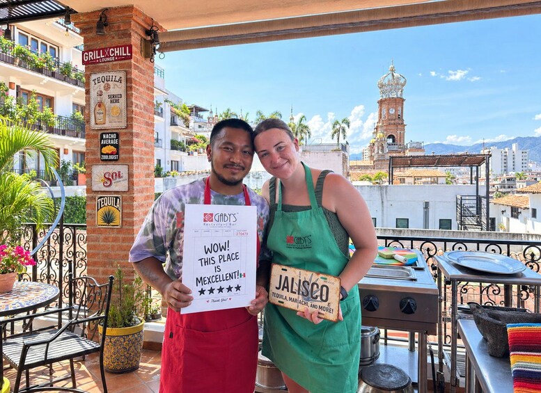 Two people holding a sign that says 'Wow! This place is Mexcellent!' with a rating of five stars. They are wearing aprons and standing on a balcony with a view of a cityscape, including a clock tower. 2
