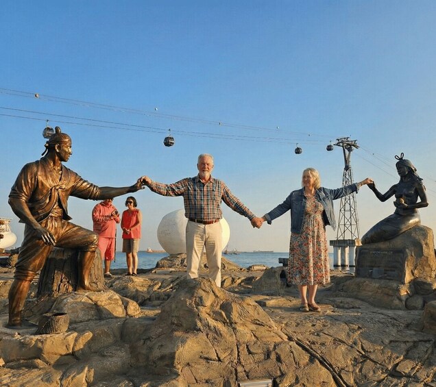 A group of people holding hands in front of a statue on a rocky beach with a cable car in the background. 3