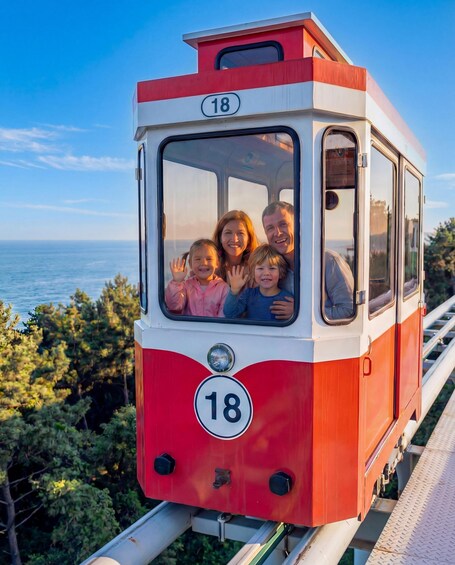 A red and white cable car with the number 18, passengers waving, and a coastal landscape in the background. 1