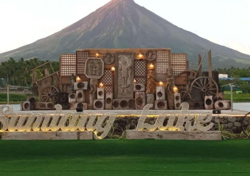 A wooden sign with 'Sundong Lake' written on it, set against a backdrop of a mountain and palm trees. 3