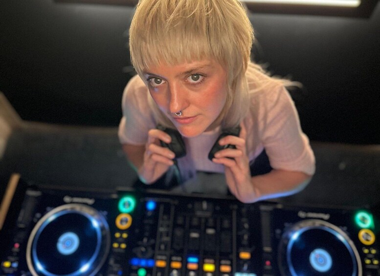 A person with blonde hair, wearing a light-colored shirt, operating DJ equipment with multiple controls and illuminated buttons. 1