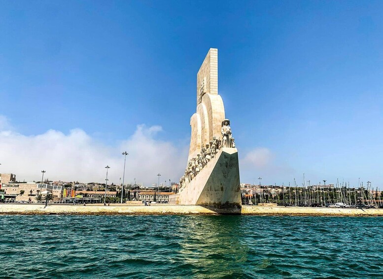 Et stort, vertikalt monument med en statue øverst, beliggende ved en vandkant med bygninger og en sandstrand i baggrunden. 2