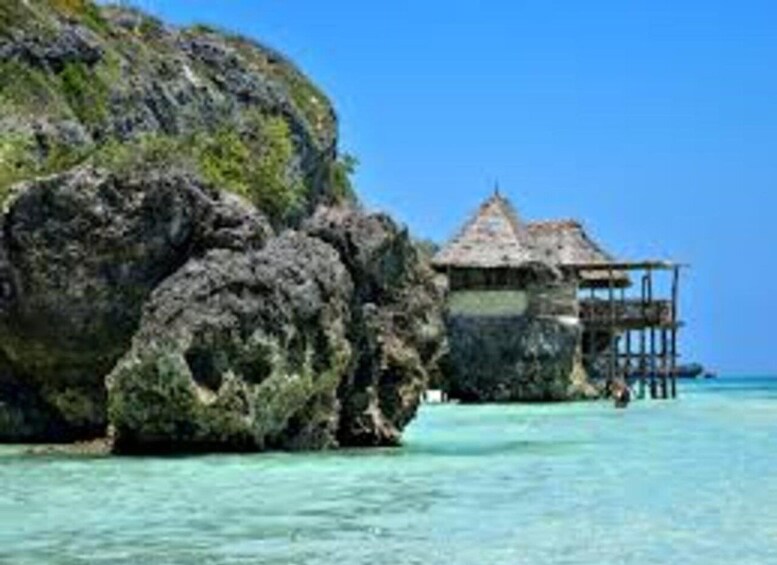 A tropical island with clear turquoise waters, a large rock formation, and a thatched-roof overwater bungalow. 5