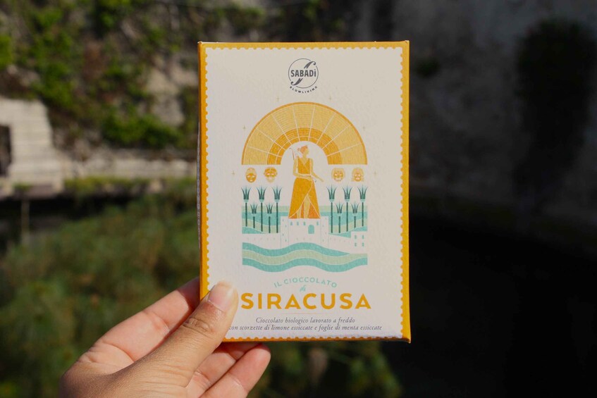 A hand holding a box of 'SIRACUSA' chocolate with a scenic background. 4