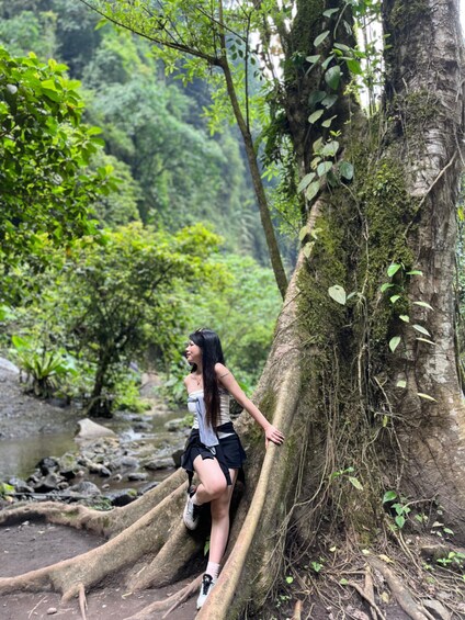 A person leaning against a tree in a forested area with a stream in the background. 2