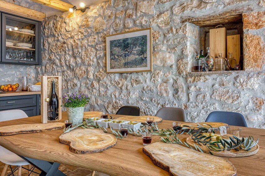 A rustic dining table set with wooden boards, olive branches, and glasses of red wine against a stone wall backdrop. 4