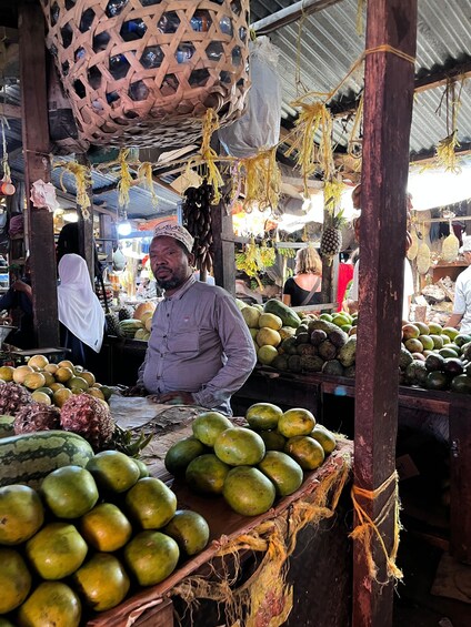 Un puesto de mercado con diversas frutas y verduras, incluyendo sandías y piñas, bajo una estructura improvisada. 2