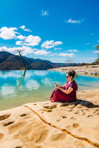 A person sitting on a sandy shore by a clear blue lake with mountains in the background. 5