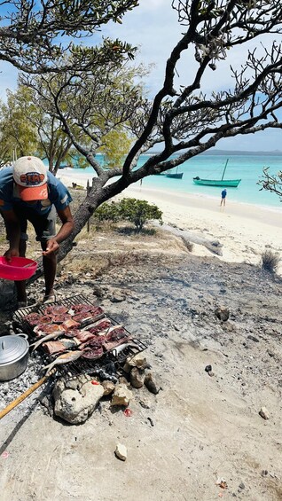 A person grilling meat on a beach with a view of the ocean and boats in the background. 3