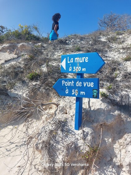 A person walking on a sandy beach with two directional signs, one pointing to 'La mure à 300 m' and the other to 'Point de vue à 58 m'. 4