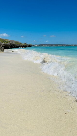 A sandy beach with gentle waves, clear blue water, and a distant rocky shoreline. 5