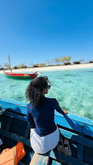 A person on a boat overlooking clear turquoise water with a beach and huts in the background. 1