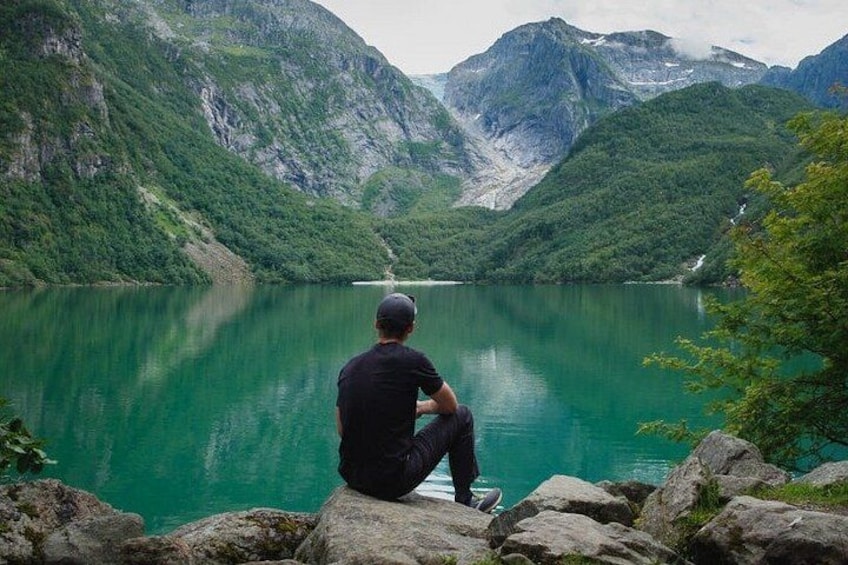 A person sitting on a rock by a turquoise lake surrounded by green mountains. 1