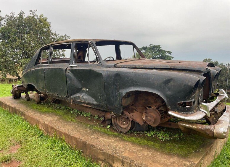 An old, rusted car on a concrete platform surrounded by grass and trees. 1