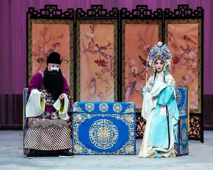 Two individuals in traditional costumes seated on ornate chairs with a backdrop of floral patterns. 3