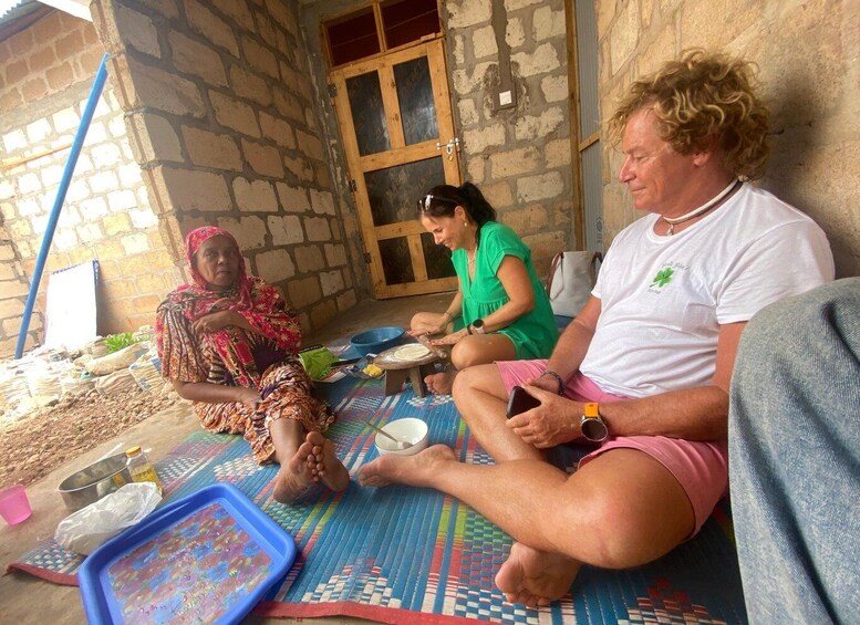 Three people sitting on a mat outside a stone building. 2