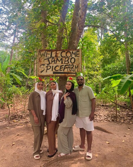 Four people standing in front of a rustic welcome sign in a lush, green forest. 1