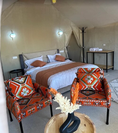 A tented accommodation with a bed, two patterned armchairs, a small table with a vase, and a coat rack. 3