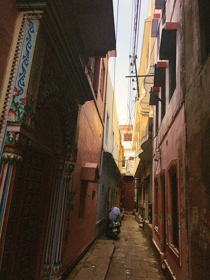 A narrow alleyway with buildings on either side, a person sitting on a motorcycle, and overhead electrical wires. 2