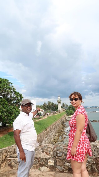 Two people standing by a stone wall overlooking a coastal area with a lighthouse in the distance. 1