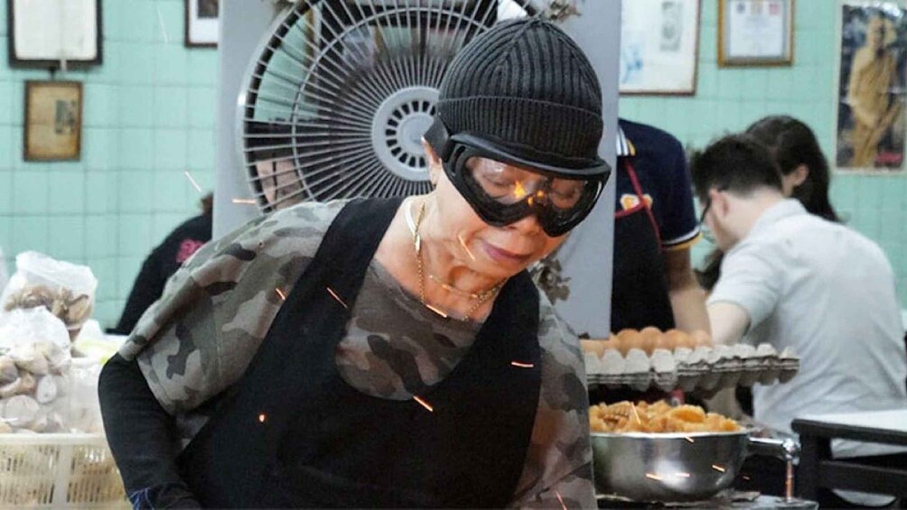 A person in a camouflage shirt and black hat working at a food stall with a fan and cooking equipment in the background. 1