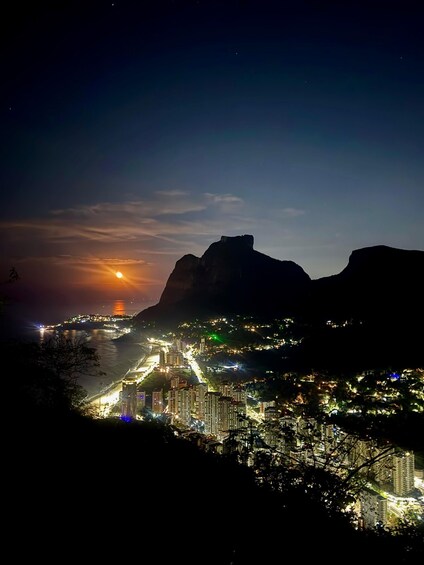 A cityscape at night with a prominent mountain range in the background and a sunset over the ocean. 5