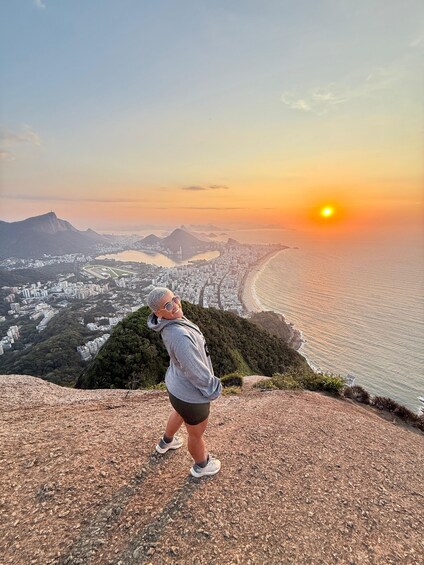 A person standing on a rocky outcrop overlooking a coastal city with a sunset in the background. 4
