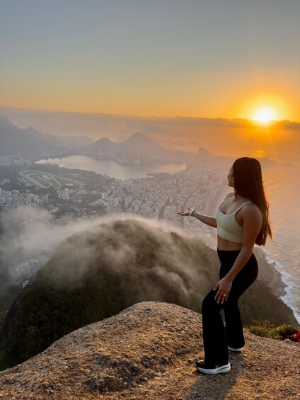 A person standing on a rocky outcrop overlooking a cityscape with a mountain in the background and the sun setting on the horizon. 3
