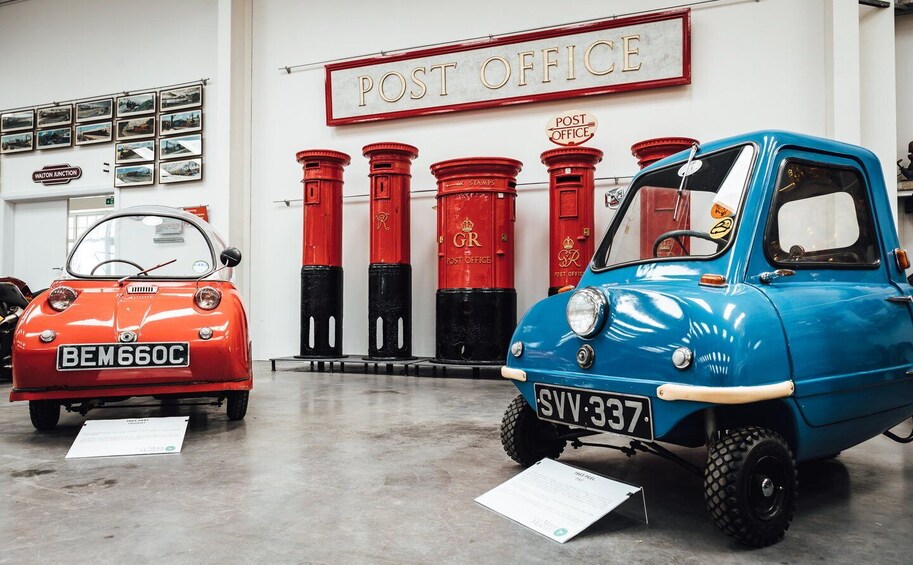 Vintage postal vehicles in a museum with a 'POST OFFICE' sign and red mailboxes. 1
