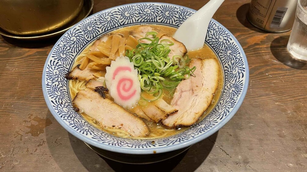 A bowl of ramen with sliced pork, green onions, and a pink swirl garnish on a patterned plate. 2