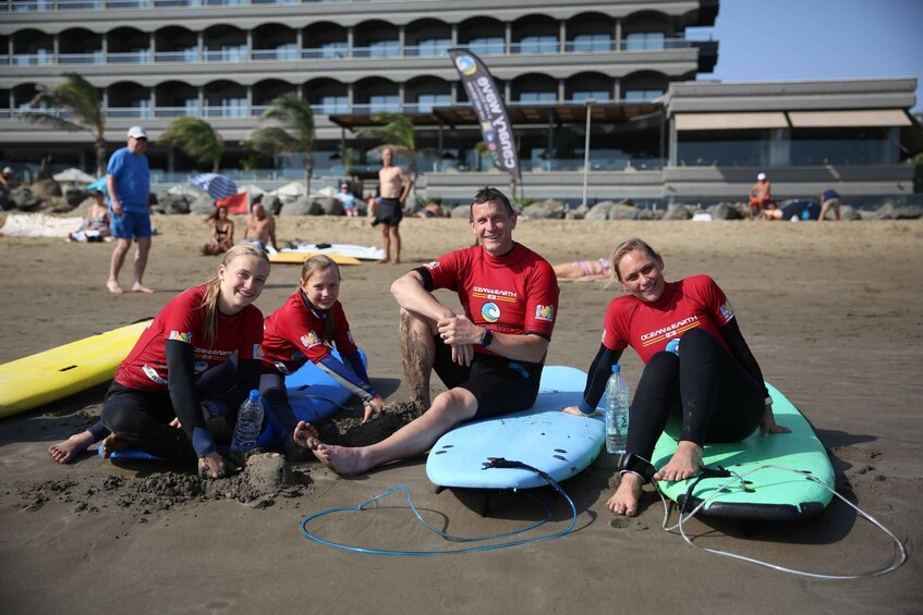 A group of people on a beach with surfboards, a building in the background, and a clear sky. 5