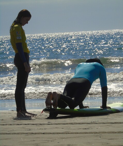 A person in a blue wetsuit lying on a surfboard on the beach, with another person standing nearby. 4