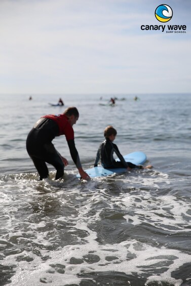 A person in a red and black wetsuit assisting another person on a blue surfboard at the beach. 4