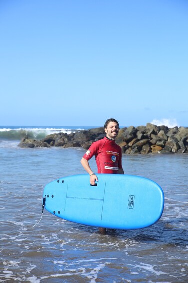 A person in a red wetsuit holding a blue surfboard with the OceanEarth logo, standing in shallow water with waves and rocks in the background. 3