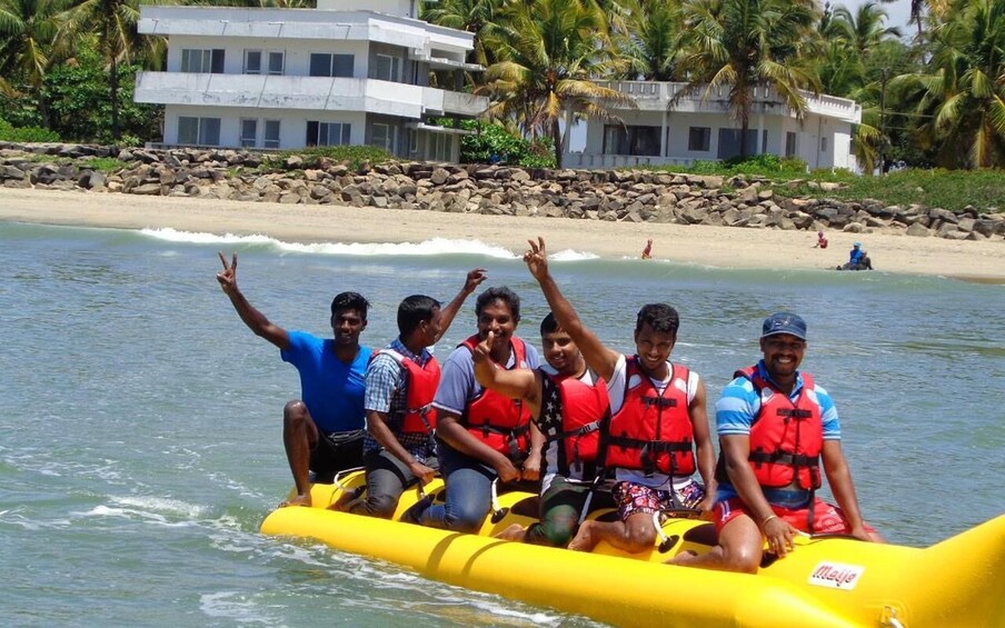A group of people in a yellow raft on calm water, wearing life jackets, with a beach and palm trees in the background. 4