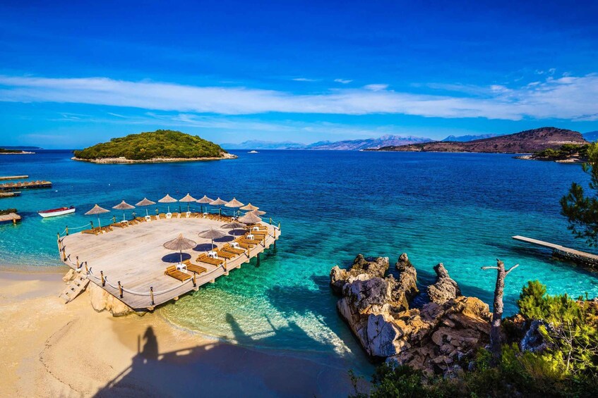 A wooden deck with umbrellas, clear turquoise waters, a sandy beach, and rocky outcrops. 1