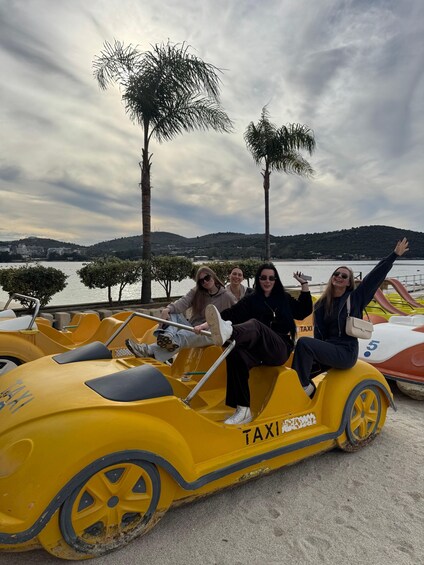 Four people in yellow taxi-shaped vehicles by a lake with palm trees and hills in the background. 4