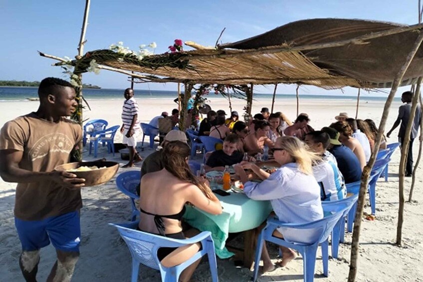 A beachside gathering with people seated at tables under a thatched structure. 1