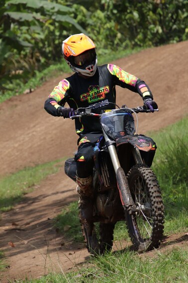 A person on a dirt bike wearing a helmet and colorful gear, riding on a dirt path surrounded by greenery. 3