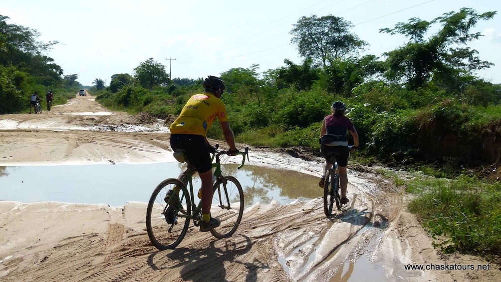 Two cyclists on a muddy trail with puddles and vegetation on the sides. 4