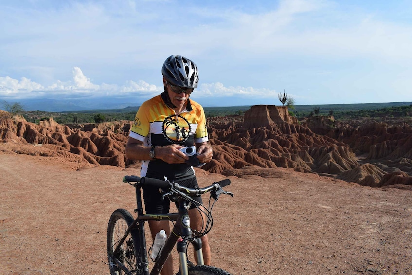 A cyclist in a yellow and black jersey, adjusting a camera on a bicycle in a desert landscape with red rock formations. 2