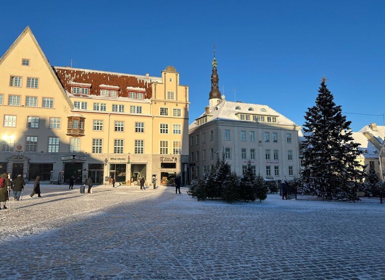 A snow-covered cobblestone square with historic buildings, a church spire, and a Christmas tree. 2