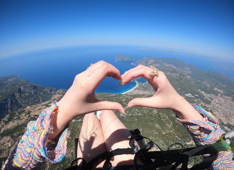 A person making a heart shape with their hands, overlooking a coastal landscape with a clear blue sea and distant islands. 2