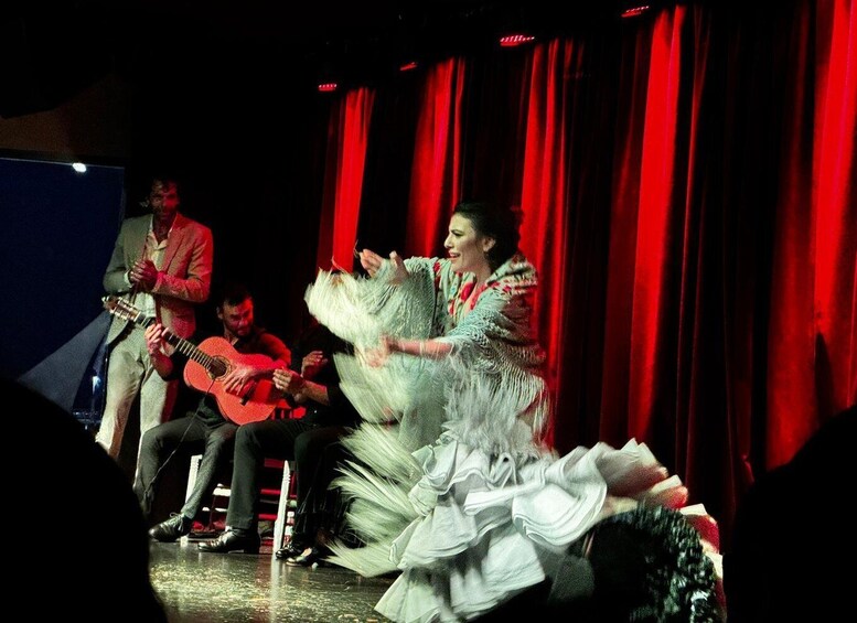 A stage performance with a dancer in a white costume, red curtains, and musicians with guitars. 1